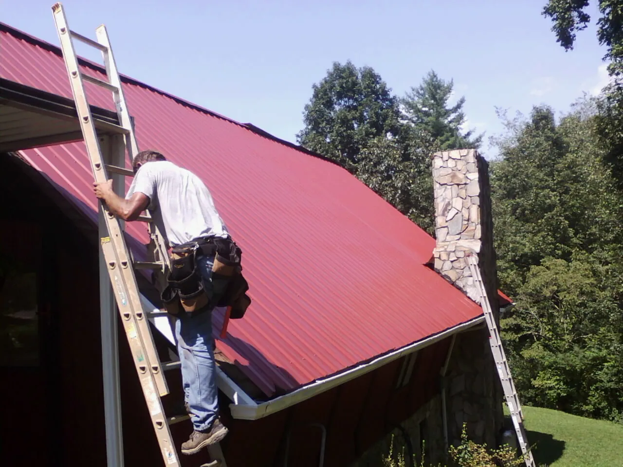 Roofer on a ladder beside a red metal roof and stone chimney — Soffit & Fascia Repair by Roofing Brothers in Richlands